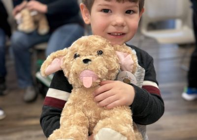 Child holding a plush tan dog toy in a room with others in the background.