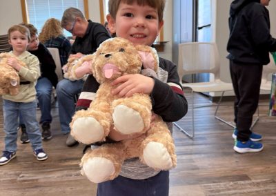 Child holding a plush stuffed animal in a room with people seated in the background.